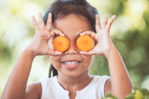 Young child holding carrot slices over their eyes and smiling outdoors